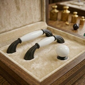 May include: Two arched cabinet handles and one spherical cabinet knob, each featuring a smooth white ceramic center section flanked by dark, ribbed metal ends, presented inside a beige velvet-lined wooden display box with gold trim.