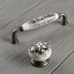 May include: A white ceramic cabinet handle and a matching round cabinet knob, both featuring a black floral pattern and an antique-style dark metal finish on the mounting hardware and base, providing a coordinated set for furniture hardware.