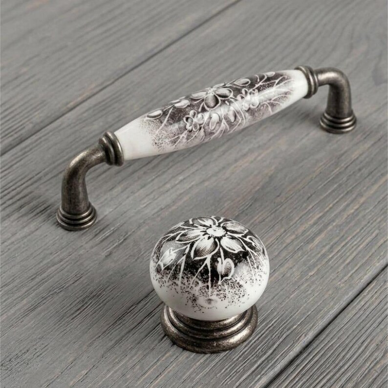 May include: A white ceramic cabinet handle and a matching round cabinet knob, both featuring a black floral pattern and an antique-style dark metal finish on the mounting hardware and base, providing a coordinated set for furniture hardware.