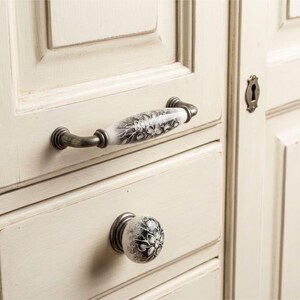 May include: A cabinet handle and a matching round knob attached to a cream-colored wooden drawer. Both pieces feature a white ceramic center with a black floral pattern and antique-style metal hardware on the ends and base.