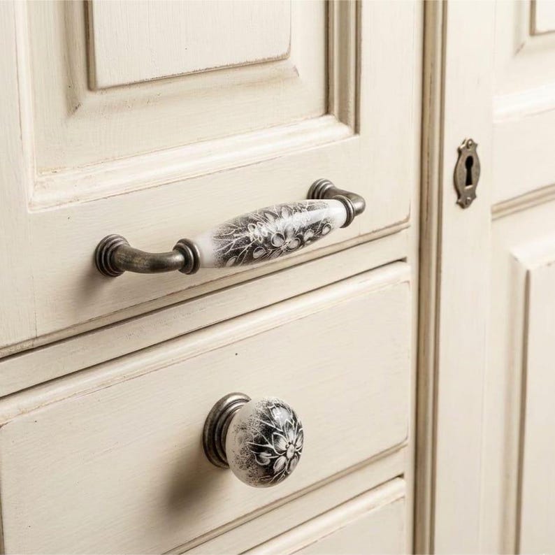 May include: A cabinet handle and a matching round knob attached to a cream-colored wooden drawer. Both pieces feature a white ceramic center with a black floral pattern and antique-style metal hardware on the ends and base.