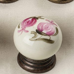May include: A round ceramic cabinet knob featuring a hand-painted floral design with two pink blossoms and green leaves on an off-white background. The knob is mounted on a circular, dark bronze-toned metal base with a tiered, ridged edge design.