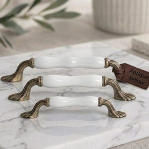 Può includere: Tre maniglie per cassetti di dimensioni diverse, ciascuna con un corpo centrale in ceramica bianca liscia e terminali in metallo color ottone anticato. Una piccola etichetta in pelle marrone sulla maniglia più grande riporta il testo Antique Yellow.