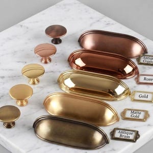 May include: A collection of metal cabinet hardware including round knobs and elongated cup pulls in various finishes such as antique copper, rose gold, polished gold, matte gold, and antique brass, each paired with a small rectangular label.