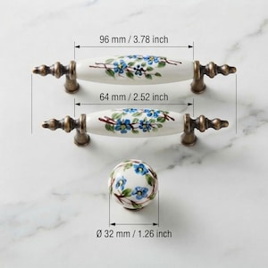 Op de afbeelding: Twee keramische ladehandgrepen en een ronde knop met een blauw bloemmotief op een witte achtergrond en antiek bronzen beslag. De handgrepen meten 96 mm en 64 mm, terwijl de knop een diameter van 32 mm heeft.