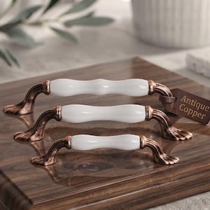 Peut inclure: Trois poignées de meuble de longueurs variées, chacune dotée d'une barre centrale en céramique blanche lisse et d'extrémités en métal cuivre antique au design cannelé en éventail. Une étiquette brune indique Antique Copper.