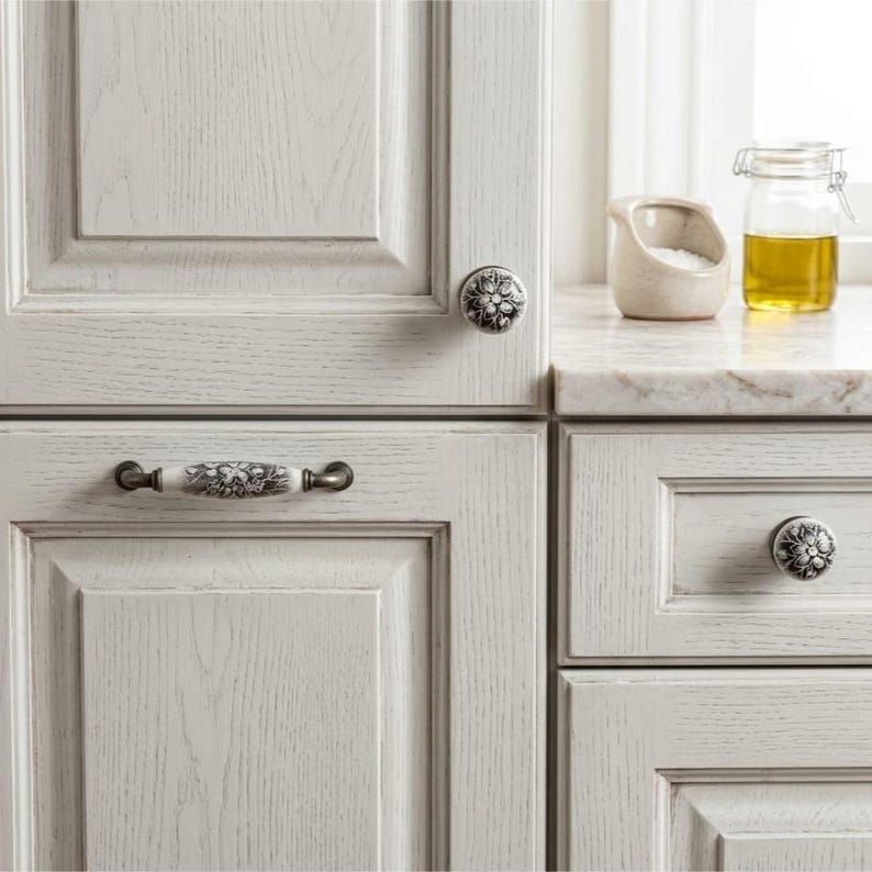 May include: Off-white wooden kitchen cabinets featuring decorative hardware. The set includes two round knobs with a black floral pattern and one horizontal handle with a matching black floral design, all mounted on light-colored wood panels.