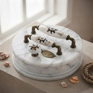 May include: Two white ceramic drawer pulls and one matching round knob, each featuring a black ship wheel design and antique brass hardware. The hardware is displayed on a white marble surface with a small oval tag labeled Antique Brass.