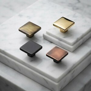May include: Four square cabinet knobs in distinct metallic finishes. One knob is polished gold, one is brushed brass, one is matte black, and one is a warm copper tone. Each knob features a flat top surface and a central mounting base.