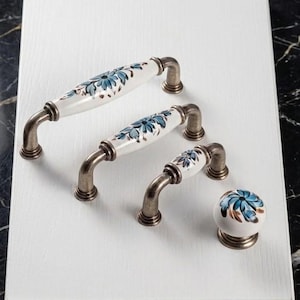 Puede incluir: Tres tiradores de armario y un pomo redondo de cerámica blanca con motivos florales azules y detalles marrones. Cada pieza cuenta con una base y estructura de metal en tono bronce antiguo con un diseño curvo y suave.