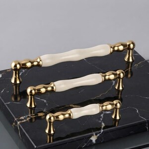 Puede incluir: Tres tiradores de armario de diferentes longitudes dispuestos en pila. Cada tirador presenta una barra central de cerámica de color crema con un perfil ondulado y suave, flanqueada por extremos de metal dorado pulido y bases de montaje redondeadas.
