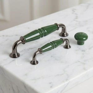 Puede incluir: Dos tiradores de armario de metal oscuro con centros de cerámica verde bosque lisos y brillantes, junto con un pomo de armario redondo de cerámica a juego. Los herrajes presentan extremos curvos y un acabado pulido que contrasta con el metal.