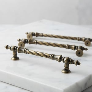 May include: Three antique brass cabinet pulls featuring a twisted rope-style center bar with decorative fluted end posts and circular mounting bases. The metal hardware has a darkened, weathered finish with visible highlights on the ridges.