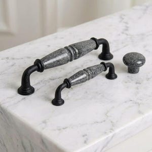 Può includere: Due maniglie per armadietti e un pomello rotondo. Ogni pezzo presenta un centro in ceramica grigia maculata e testurizzata con finitura lucida, accentuato da ferramenta in metallo nero opaco alle estremità e alla base delle maniglie.