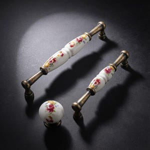 May include: Two drawer pulls and one round cabinet knob featuring white ceramic centers with a floral pattern of red roses and yellow accents, finished with antique brass metal hardware on the ends and base for a classic furniture look.