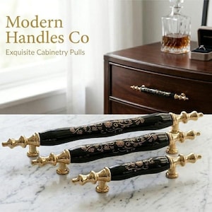 May include: Three cabinet pulls of varying lengths featuring a glossy black cylindrical center with gold floral patterns and gold metal ends. The text Modern Handles Co Exquisite Cabinetry Pulls is visible in the upper left corner of the image.