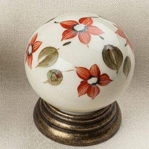 May include: A round ceramic cabinet knob featuring a cream-colored base with hand-painted orange flowers and green leaves. The knob is mounted on a circular, dark bronze-toned metal base with a textured, weathered finish and tiered detailing.