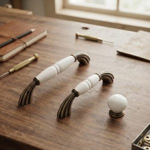 May include: Two arched cabinet pulls and one round cabinet knob. Each pull features a smooth white ceramic centre section with ribbed, antique brass-toned metal ends. The round knob is solid white ceramic with an antique brass-toned metal base.