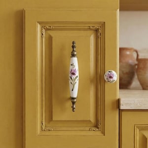 May include: A mustard yellow cabinet door featuring a vertical ceramic handle and a matching round knob. Both hardware pieces are white with a hand-painted purple flower and green leaves. The cabinet door has a raised rectangular frame design.