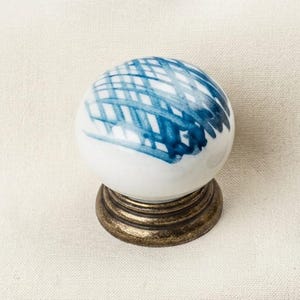 May include: A round white ceramic cabinet knob featuring a hand-painted blue cross-hatch pattern on its top surface. The knob is mounted on a circular, dark bronze-toned metal base with a tiered, ridged design that supports the spherical top.
