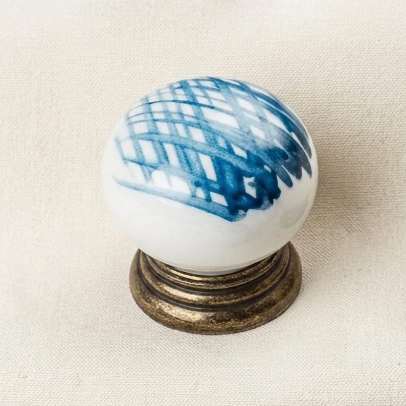 May include: A round white ceramic cabinet knob featuring a hand-painted blue cross-hatch pattern on its top surface. The knob is mounted on a circular, dark bronze-toned metal base with a tiered, ridged design that supports the spherical top.