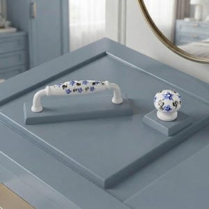 May include: A white ceramic cabinet handle and a matching round cabinet knob, both featuring a hand-painted blue floral pattern with green leaves. The hardware is mounted on a smooth, matte blue surface, showcasing a clean and functional design.
