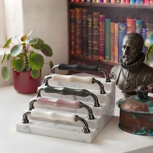 May include: A tiered white marble display stand holding four cabinet pulls. The pulls feature dark metallic hardware with central ceramic grips in matte black, cream, sage green, dusty rose, and white, arranged in a vertical staircase formation.