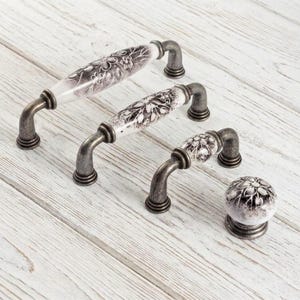 May include: A collection of three arched cabinet pulls and one round cabinet knob. Each piece features a white ceramic center with a dark gray floral pattern and is finished with weathered, dark metallic hardware at the mounting points.