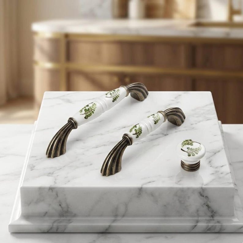 May include: Two arched cabinet handles and one round cabinet knob, each featuring a white ceramic center with a green fern leaf pattern and antique bronze-toned metal ends with a ribbed, fluted design on the mounting hardware.