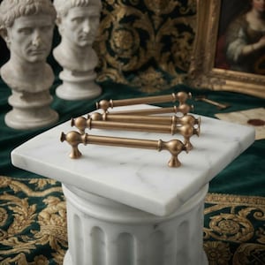 Może przedstawiać: Trzy cylindryczne mosiężne uchwyty meblowe z zaokrąglonymi końcami i gładkim, matowym wykończeniem metalicznym. Każdy uchwyt posiada centralny pręt wsparty na dwóch małych, rozszerzających się podstawach, ułożone równolegle na białej marmurowej powierzchni.