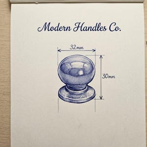 May include: A blue ink sketch on white paper featuring a round cabinet knob with a circular base. The text Modern Handles Co. is printed above the knob. Dimension lines indicate a width of 32mm and a height of 30mm for the hardware component.