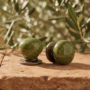 May include: Two round cabinet knobs featuring a glossy, moss-green ceramic surface with a fine, dark crackle glaze pattern. Each knob is mounted on a circular, weathered bronze-toned metal base, providing a contrast to the smooth, rounded top.