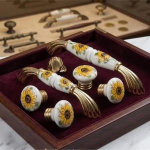 Può includere: Un set di ferramenta per mobili composto da due maniglie arcuate e quattro pomelli rotondi. Ogni pezzo è realizzato in ceramica bianca con disegni di girasoli gialli dipinti a mano e terminali in metallo dorato, in una scatola di legno.
