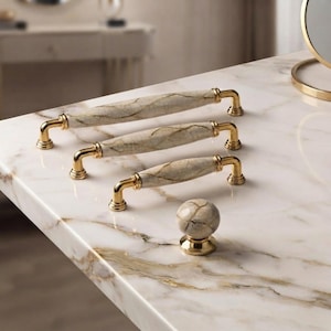 May include: Three cabinet pulls and one round cabinet knob featuring a beige and grey marbled stone center with gold-toned metal hardware. The pulls vary in length, each showcasing a smooth, cylindrical stone body with gold-finished end caps.