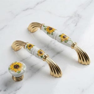 May include: Two arched cabinet handles and one round cabinet knob made of white ceramic with yellow sunflower patterns and gold-toned metal ends. The handles feature a ribbed design on the metallic sections, while the knob has a smooth finish.