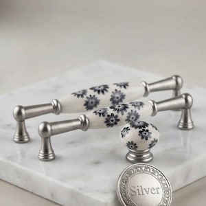 May include: Two cabinet pulls and one cabinet knob featuring a white ceramic center with a black floral pattern and brushed silver metal ends. A round silver metal disc with the word Silver embossed in the center is positioned in the foreground.