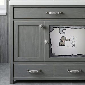 May include: A gray wooden bathroom vanity featuring one top drawer, two lower drawers, and a cabinet door. The cabinet door displays a hand-drawn illustration of an elephant blowing bubbles under a crescent moon. The unit has white floral knobs.
