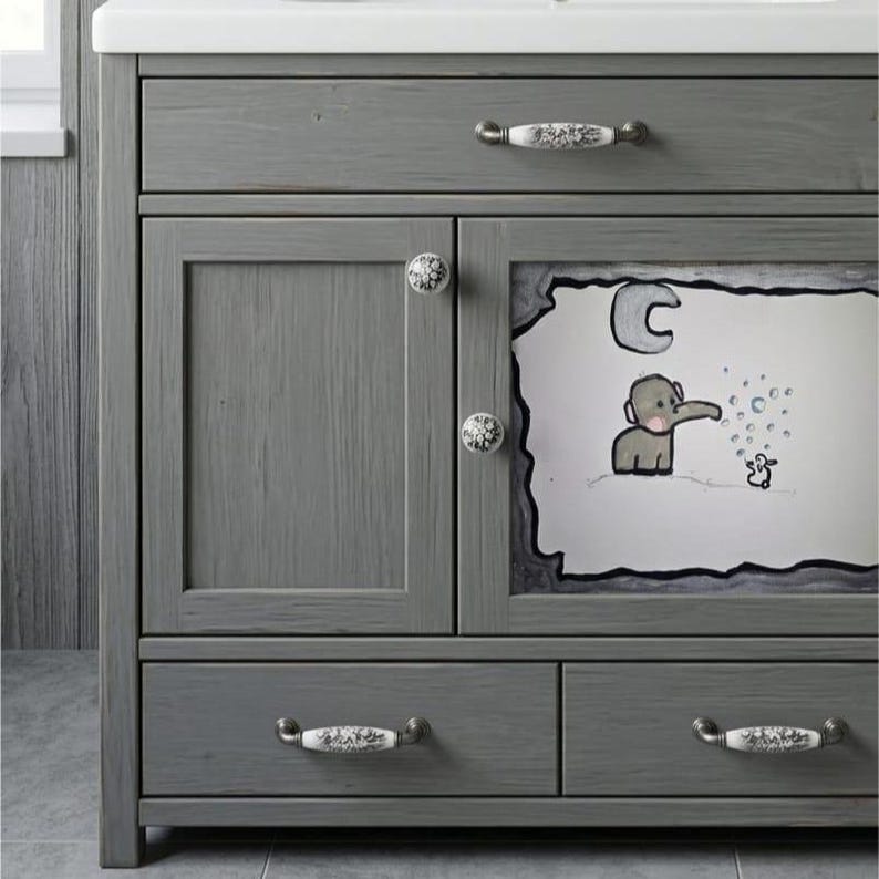 May include: A gray wooden bathroom vanity featuring one top drawer, two lower drawers, and a cabinet door. The cabinet door displays a hand-drawn illustration of an elephant blowing bubbles under a crescent moon. The unit has white floral knobs.