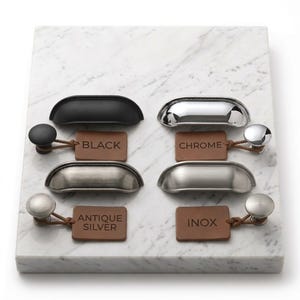 May include: A collection of four metal drawer pulls and four matching round knobs in black, chrome, antique silver, and inox finishes. Each piece is paired with a brown leather tag labeled with its respective color name in black text.