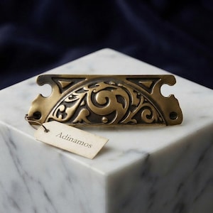 May include: A semi-circular brass drawer pull featuring an intricate embossed scrollwork pattern and two screw holes on the ends. A small, rectangular off-white paper tag is attached to the left side with a string, displaying the text Adinamos.