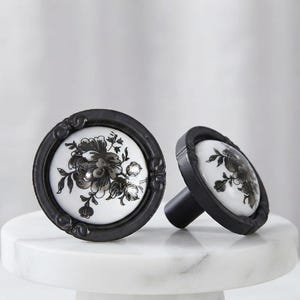 May include: Two round cabinet knobs featuring a white ceramic centre with a black floral print. Each knob is encased in a dark matte black metal frame with decorative scrollwork details along the circular edge and a cylindrical mounting post.