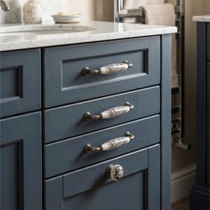 May include: A dark blue wooden bathroom vanity cabinet featuring three horizontal drawers and one bottom cabinet door. Each drawer is fitted with a white ceramic pull handle with a floral pattern and bronze-toned metal ends.