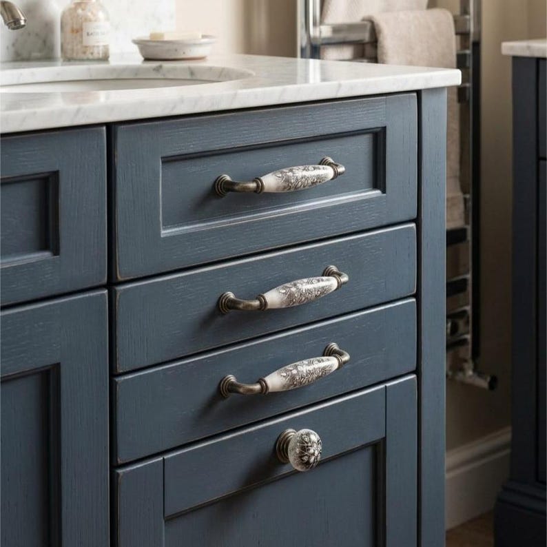 May include: A dark blue wooden bathroom vanity cabinet featuring three horizontal drawers and one bottom cabinet door. Each drawer is fitted with a white ceramic pull handle with a floral pattern and bronze-toned metal ends.