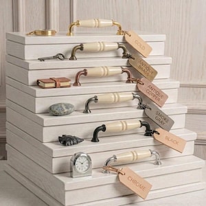 May include: A stack of six white wooden blocks, each featuring a cabinet handle with a cream-colored ceramic grip. The handles have finishes labeled Gold, Antique Brass, Antique Copper, Smoke Gray, Black, and Chrome on attached tags.