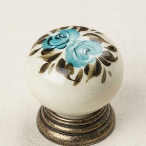 May include: A round ceramic cabinet knob with a cream-colored base, featuring hand-painted teal blue roses and dark brown leaves on the top surface. The knob is mounted on a circular, antique-style bronze metal base with a tiered, ridged design.