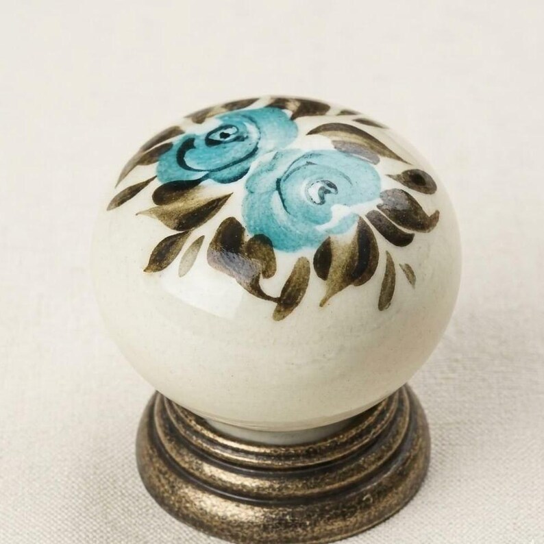 May include: A round ceramic cabinet knob with a cream-colored base, featuring hand-painted teal blue roses and dark brown leaves on the top surface. The knob is mounted on a circular, antique-style bronze metal base with a tiered, ridged design.