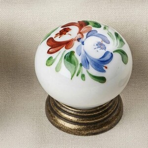 May include: A round white ceramic cabinet knob featuring a hand-painted floral design with blue, red, and green brushstrokes. The knob is mounted on a circular, textured, antique brass-toned metal base that provides a sturdy foundation for the piece.