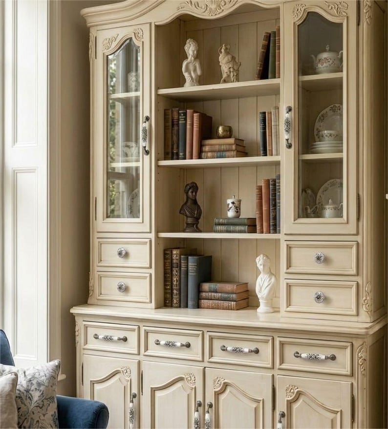 May include: A cream-colored wooden hutch with glass-paneled upper doors, open central shelving, and lower cabinets with drawers. The unit features decorative molding, ceramic hardware, and displays various books, small sculptures, and dishware.