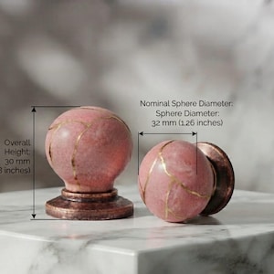 Puede incluir: Dos pomos esféricos de color rosa para armarios con vetas doradas y una base metálica oscura. La altura total es de 30 mm y el diámetro nominal de la esfera es de 32 mm, según indican las etiquetas de texto visibles en la imagen.