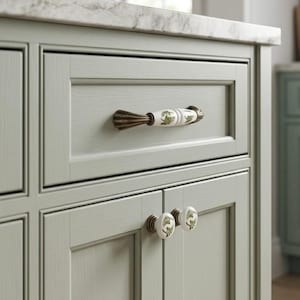 May include: A sage green cabinet featuring a single drawer with a horizontal white ceramic handle decorated with green fern patterns, and two lower cabinet doors each fitted with a round white ceramic knob featuring matching green fern designs.